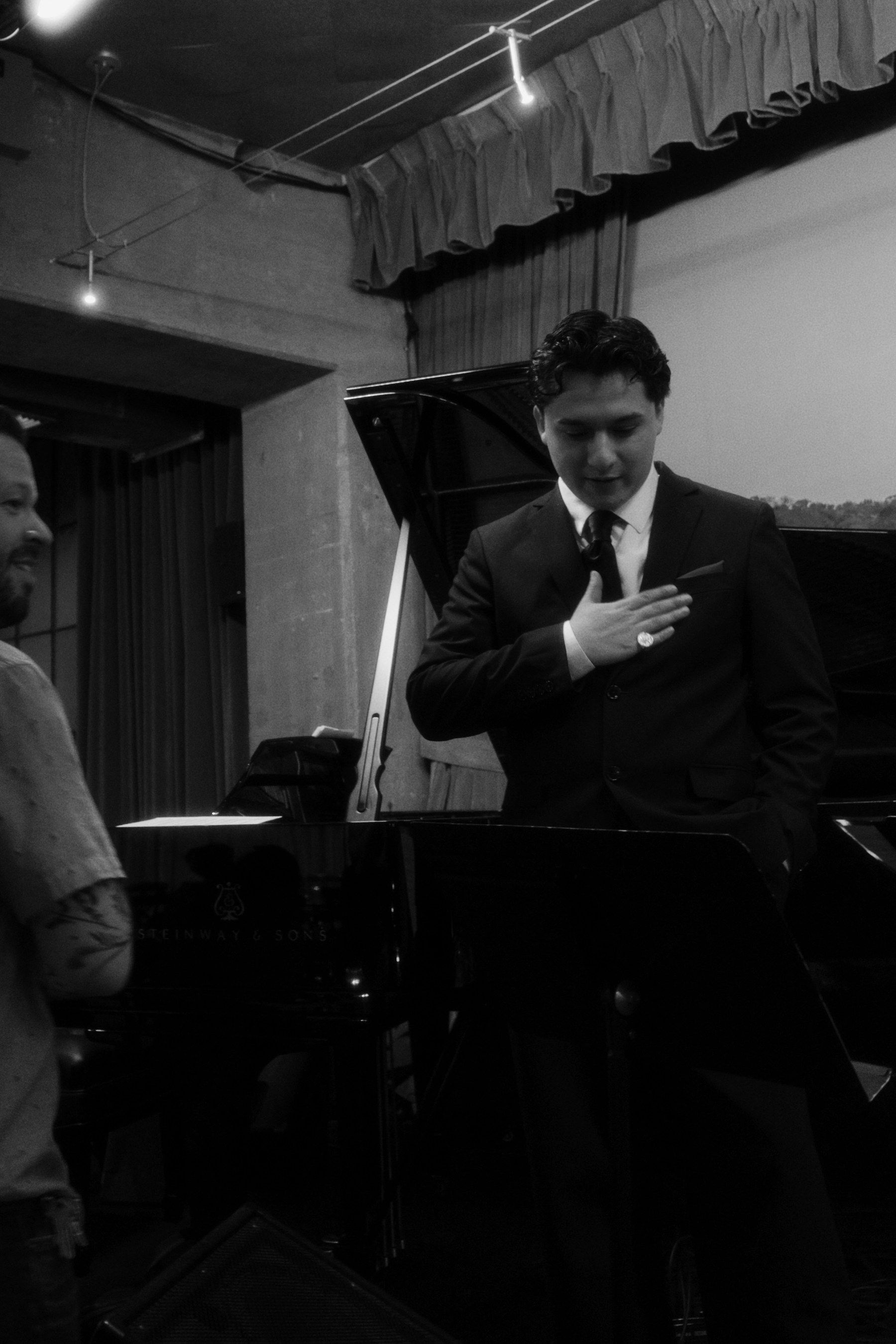 Diego on stage in a suit and tie, hand over his heart, beside a grand piano