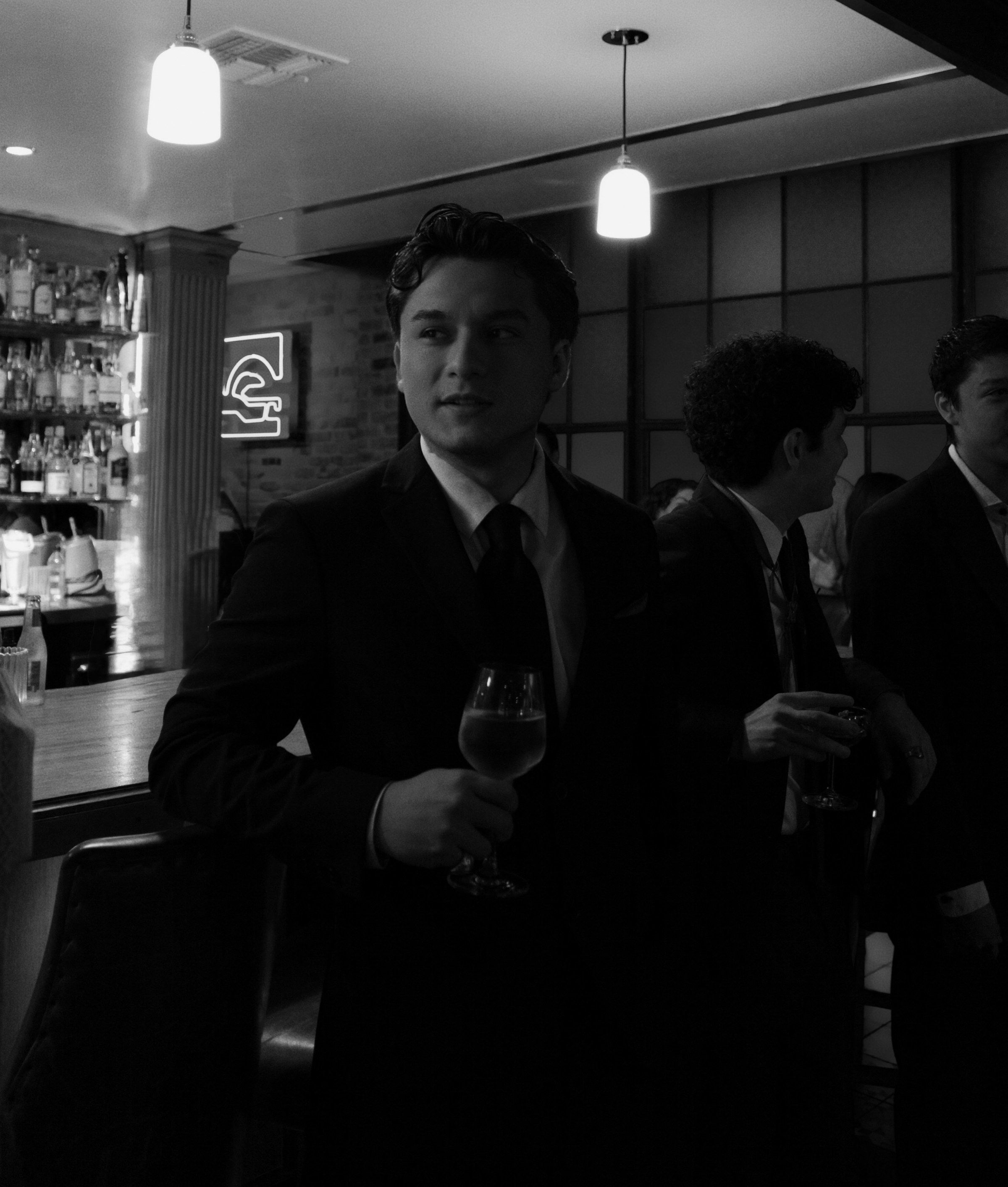Diego in a dark suit at a cocktail bar, glass in hand, pendant lights glowing overhead